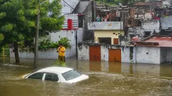 <p>A man stands near a partially submerged vehicle due to...- India TV Hindi